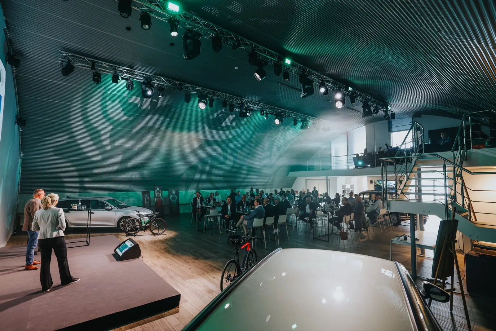 Fahrzeugpräsentation im Hangar: Moderatoren auf der Bühne, Publikum und grüne Lichtmuster unter dem Bogendach